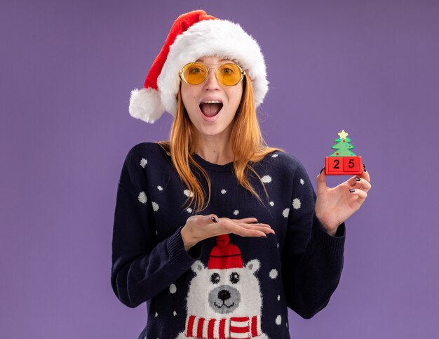 Surprised young beautiful girl wearing christmas sweater and hat with glasses holding and points with hand at christmas toy isolated on purple background