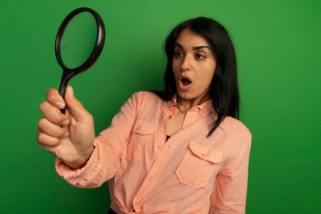 Surprised young beautiful girl wearing pink t-shirt holding and looking at magnifier isolated on green wall