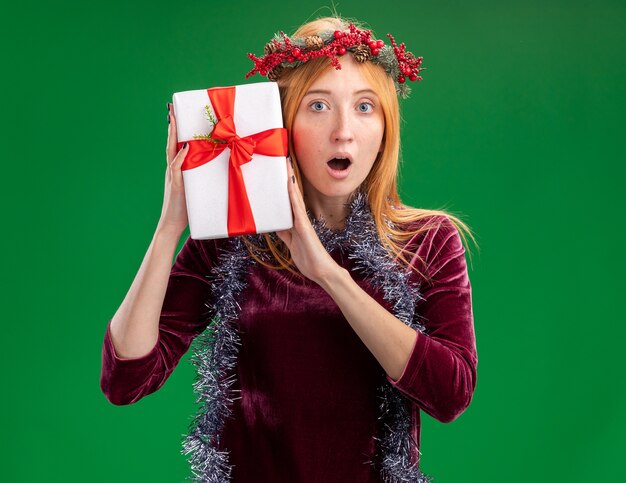 Surprised young beautiful girl wearing red dress with wreath and garland on neck holding gift box around face isolated on green background