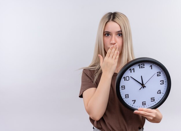 Surprised young blonde girl holding clock with hand on mouth on isolated white wall with copy space