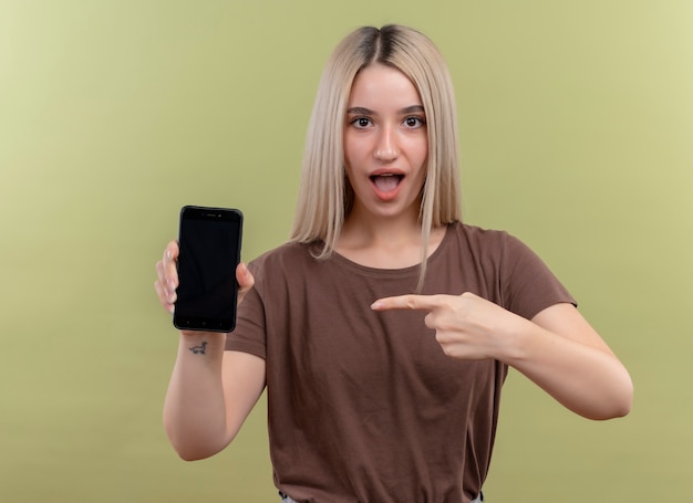 Surprised young blonde girl holding mobile phone and pointing at it on isolated green wall