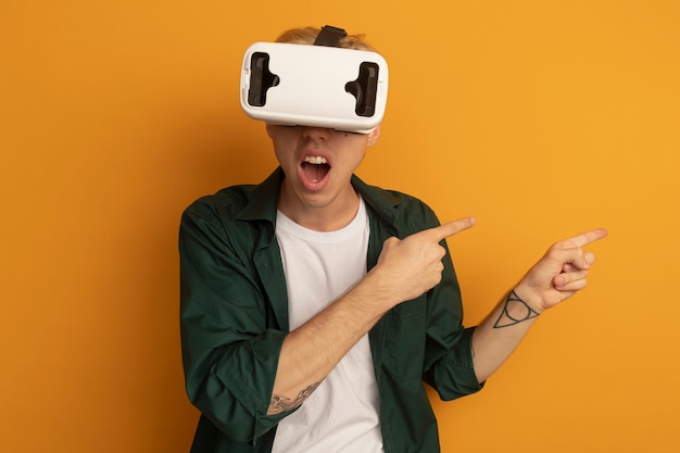 Surprised young blonde guy wearing green t-shirt and vr headset points at side