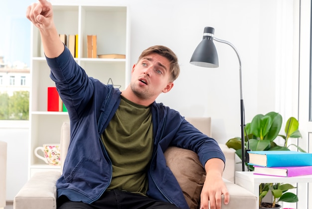 Surprised young blonde handsome man sits on armchair looking and pointing at side