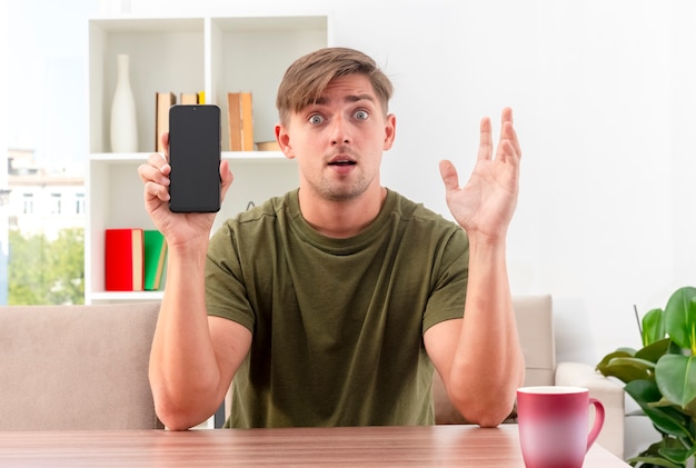 Free photo surprised young blonde handsome man sits at table with cup holding phone and raising hand inside living room