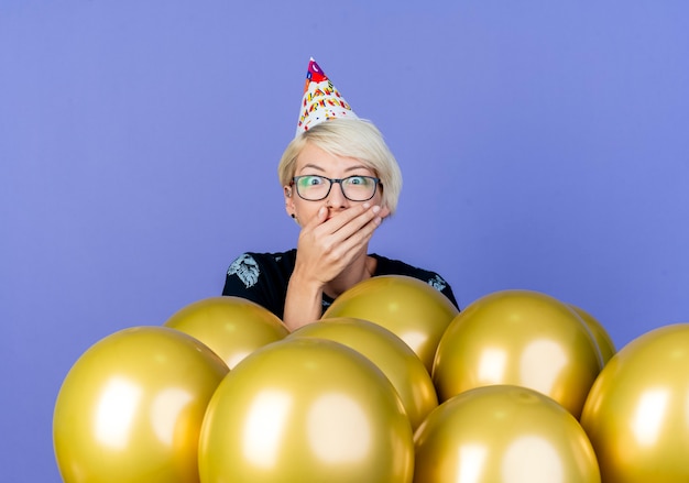 Free photo surprised young blonde party girl wearing glasses and birthday cap standing behind balloons keeping hand on mouth isolated on purple background