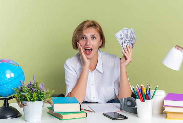 Surprised young blonde student girl sitting at desk with school tools keeping hand on face holding money 