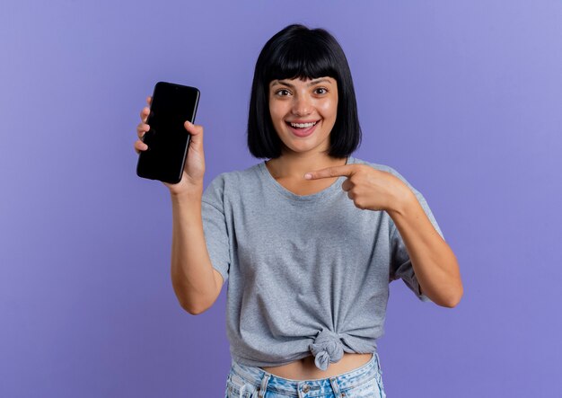 Surprised young brunette caucasian woman holds and points at phone isolated on purple background with copy space