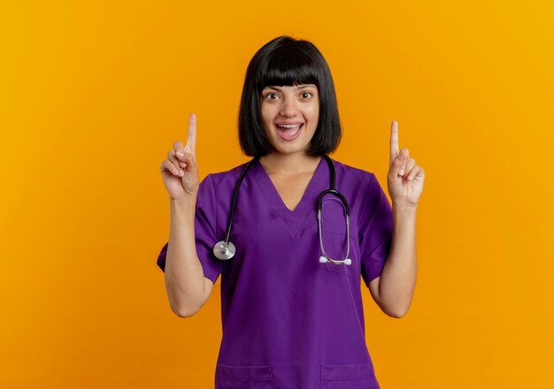 Surprised young brunette female doctor in uniform with stethoscope points up with two hands 