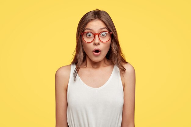 surprised young brunette with glasses posing against the yellow wall