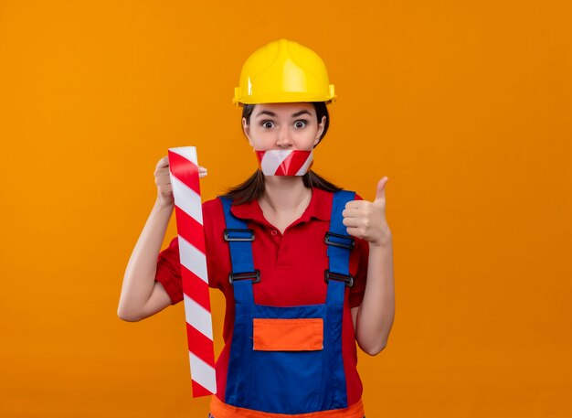 Surprised young builder girl mouth sealed with warning tape holds tape and thumbs up on isolated orange background with copy space