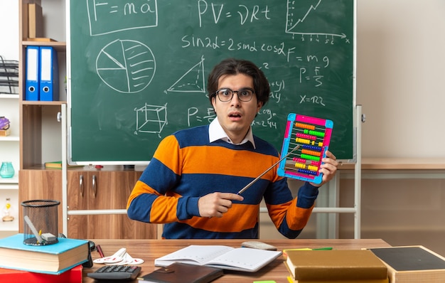 surprised young caucasian geometry teacher wearing glasses sitting at desk with school supplies in classroom showing abacus pointing at it with pointer stick looking at front