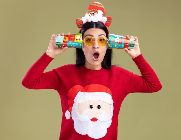 Free photo surprised young caucasian girl wearing santa claus headband and sweater with glasses holding plastic christmas cups next to ears listening to conversations  isolated on olive green wall