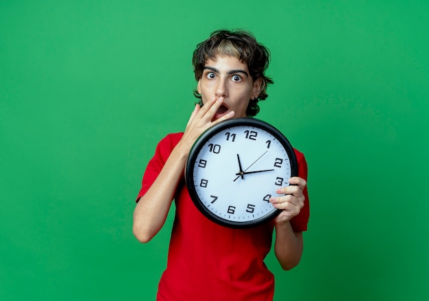 Surprised young caucasian girl with pixie haircut holding clock with hand on mouth isolated on green background with copy space