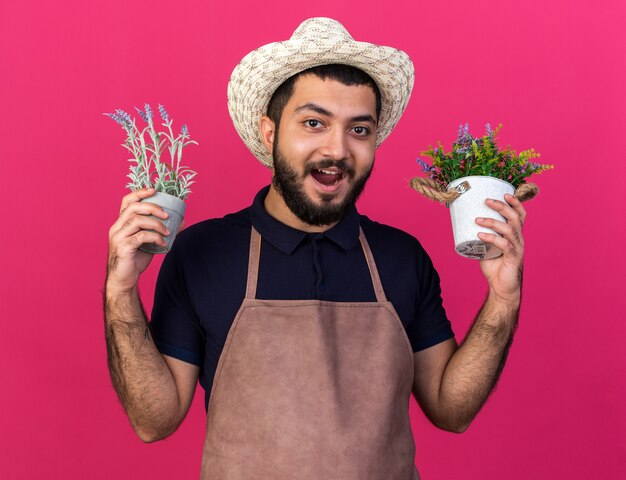 surprised young caucasian male gardener wearing gardening hat holding flowerpots isolated on pink wall with copy space