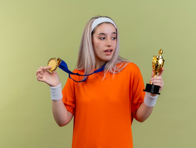 Free photo surprised young caucasian sporty girl with braces wearing headband and wristbands holds gold medal and looks at winner cup