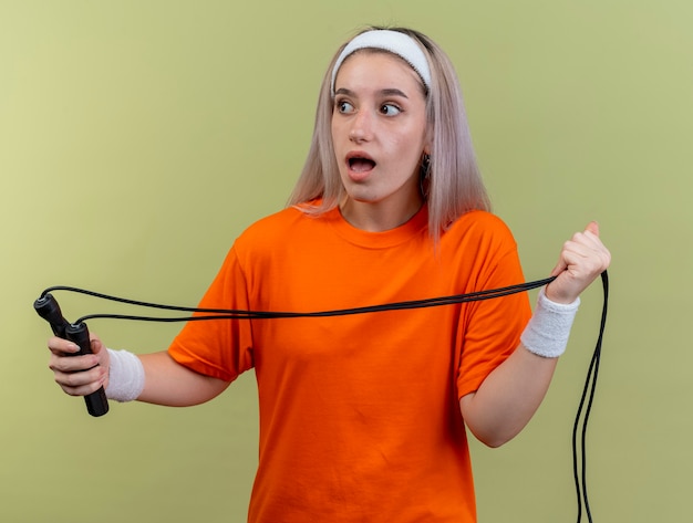 Free photo surprised young caucasian sporty girl with braces wearing headband and wristbands holds jumping rope looking at side