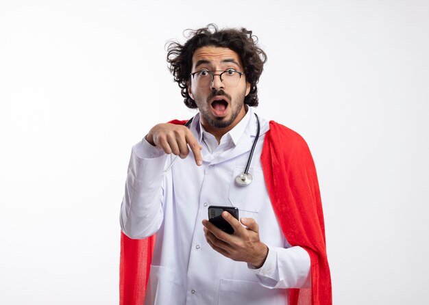 Surprised young caucasian superhero man in optical glasses wearing doctor uniform with red cloak and with stethoscope around neck holds and points at phone 