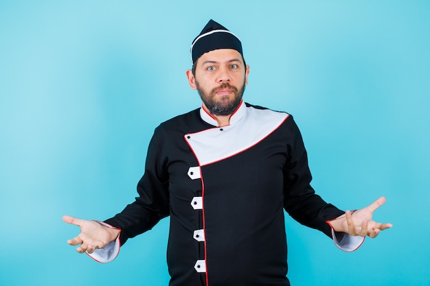 Free photo surprised young chef is looking at camera by opening wide his arms on blue background