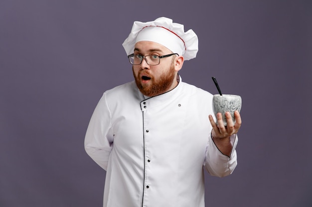 Surprised young chef wearing glasses uniform and cap showing bowl with spoon in it looking at camera while keeping hand behind back isolated on purple background