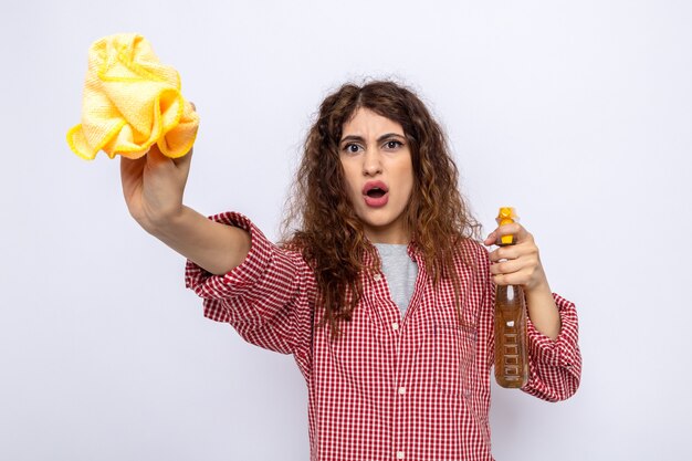 Surprised young cleaning woman holding out cleaning agent with rag