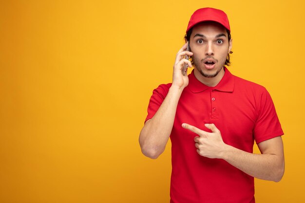 surprised young delivery man wearing uniform and cap looking at camera pointing to side isolated on yellow background with copy space