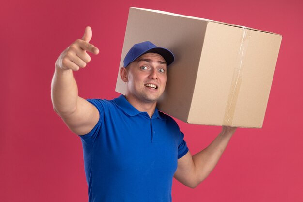 Surprised young delivery man wearing uniform with cap holding big box on shoulder and showing you gesture isolated on pink wall