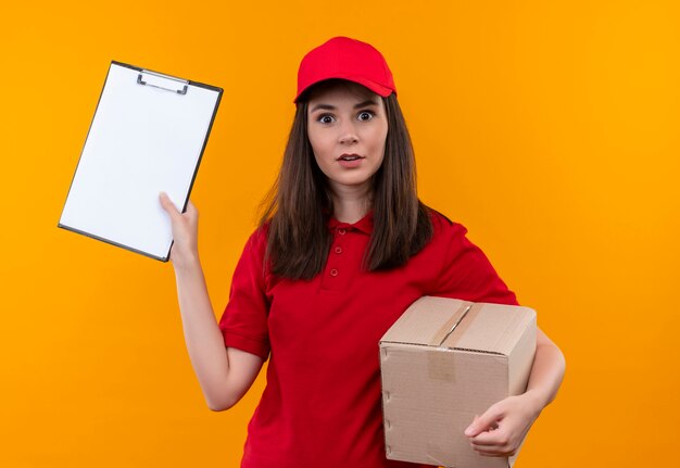 Surprised young delivery woman wearing red t-shirt in red cap holding a box and clipboard on isolated yellow wall