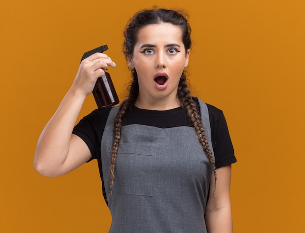 Surprised young female barber in uniform holding spray bottle isolated on orange wall