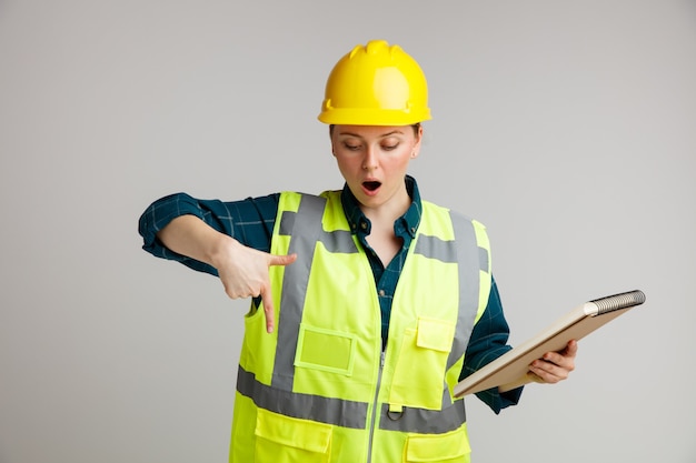 Surprised young female construction worker wearing safety helmet and safety vest holding notepad looking and pointing down 