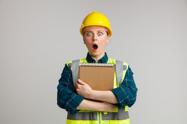 Surprised young female construction worker wearing safety helmet and safety vest hugging notepad 