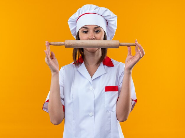 Surprised young female cook wearing chef uniform holding and looking at rolling pin isolated on orange background