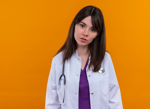 Surprised young female doctor in medical robe with stethoscope looks at camera on isolated orange background with copy space