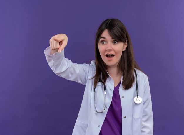 Surprised young female doctor in medical robe with stethoscope points to the side on isolated violet background with copy space