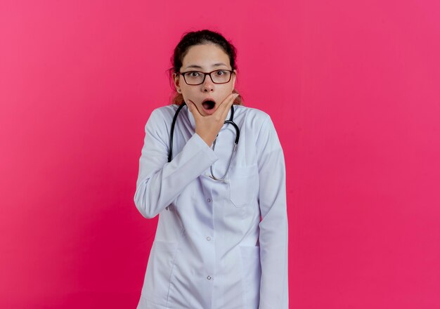 Surprised young female doctor wearing medical robe and stethoscope and glasses  holding chin isolated on pink wall with copy space