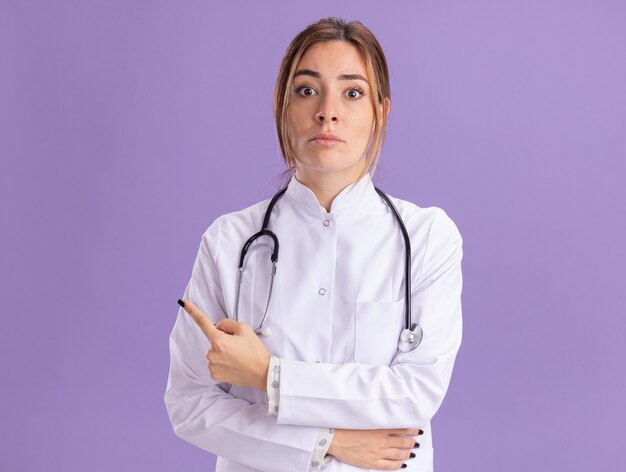 Free photo surprised young female doctor wearing medical robe with stethoscope points at side isolated on purple wall with copy space