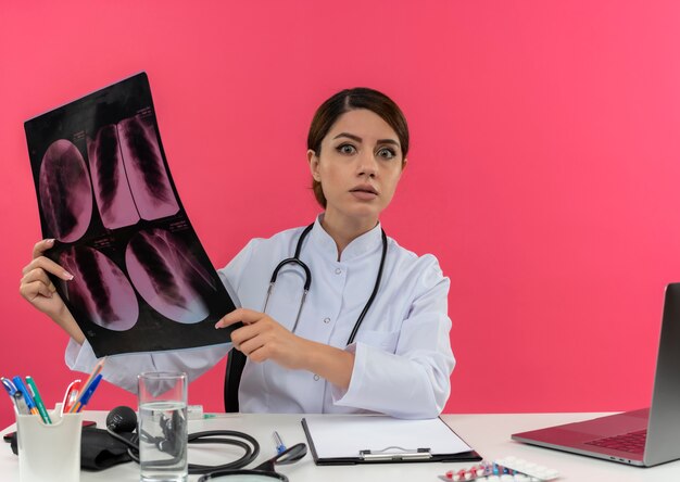 Surprised young female doctor wearing medical robe with stethoscope sitting at desk work on computer with medical tools holding x-ray with copy space