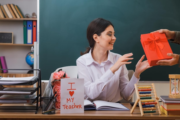 surprised young female teacher received gift box sitting at desk with school tools in classroom