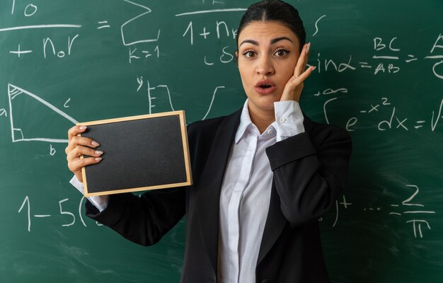 Surprised young female teacher standing in front blackboard holding mini blackboard in classroom