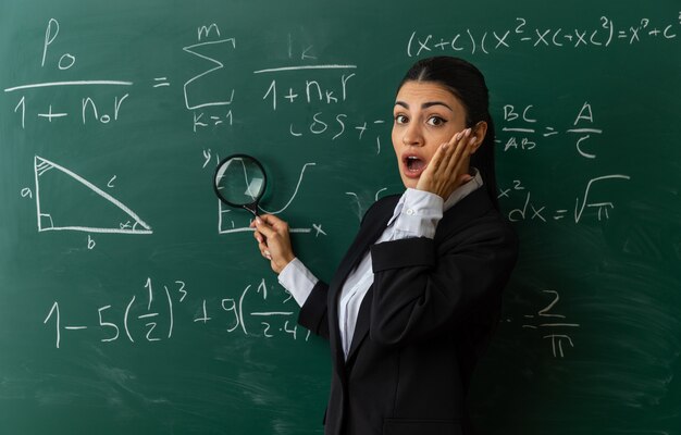 surprised young female teacher standing in front blackboard putting hand on cheek in classroom