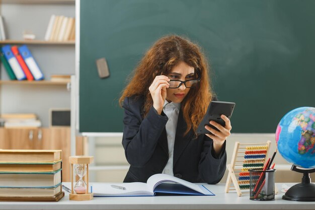 surprised young female teacher wearing glasses holding and looking at calculator sitting at desk with school tools in classroom