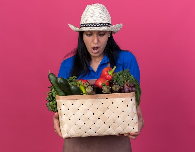 Free photo surprised young gardener woman wearing uniform and hat holding and looking at basket of vegetables isolated on pink wall with copy space