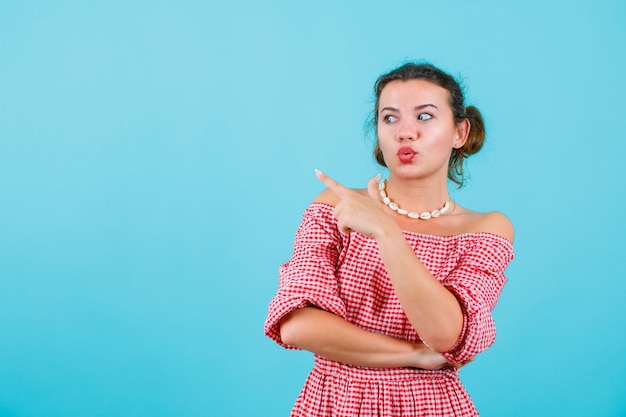 Surprised young girl is looking and pointing left with forefinger on blue background