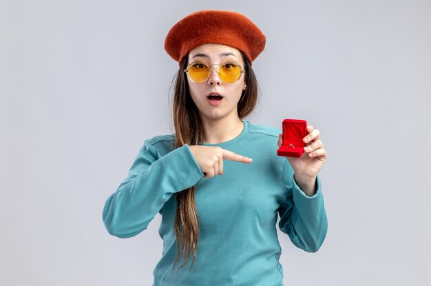 Free photo surprised young girl on valentines day wearing hat with glasses holding and points at wedding ring isolated on white background