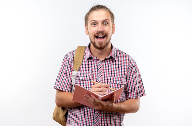 Surprised young guy student wearing backpack writing something on notebook isolated on white wall