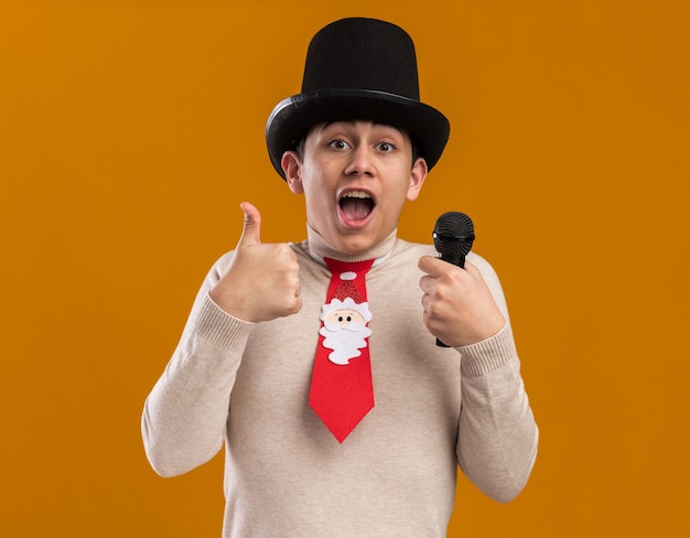 Surprised young guy wearing hat with christmas tie holding microphone showing thumb up isolated on yellow wall