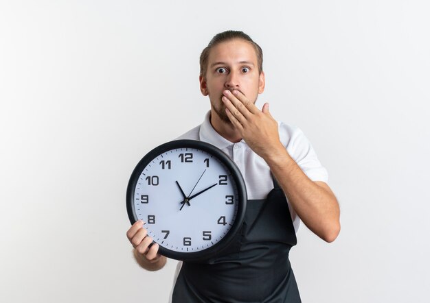 Surprised young handsome barber wearing uniform holding clock and putting hand on mouth isolated on white wall