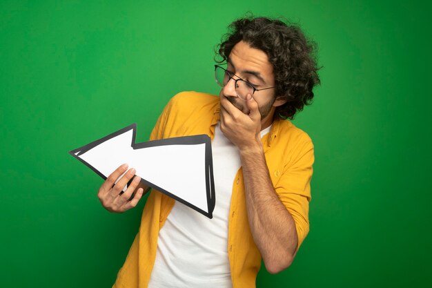 Surprised young handsome caucasian man wearing glasses holding arrow mark that is pointing at side looking at it keeping hand on mouth isolated on green background with copy space