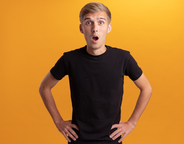 Free Photo surprised young handsome guy wearing black shirt putting hands on hip isolated on yellow wall