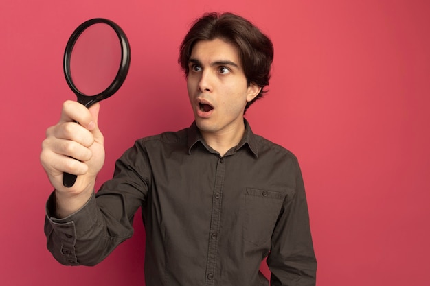 Surprised young handsome guy wearing black t-shirt holding and looking at magnifier isolated on pink wall