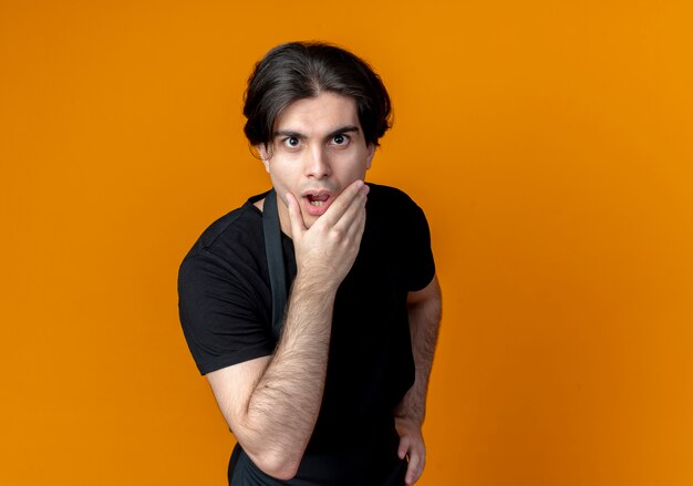 Surprised young handsome male barber in uniform grabbed chin isolated on orange background with copy space